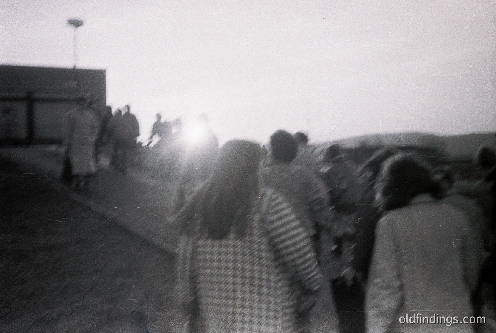 Blurred black-and-white street scene with dense crowd under low-angle sunlight. Mid-20th century urban setting, likely post-war European city. Distinctive checkered coat and lampposts suggest 1950s-60s fashion and infrastructure.