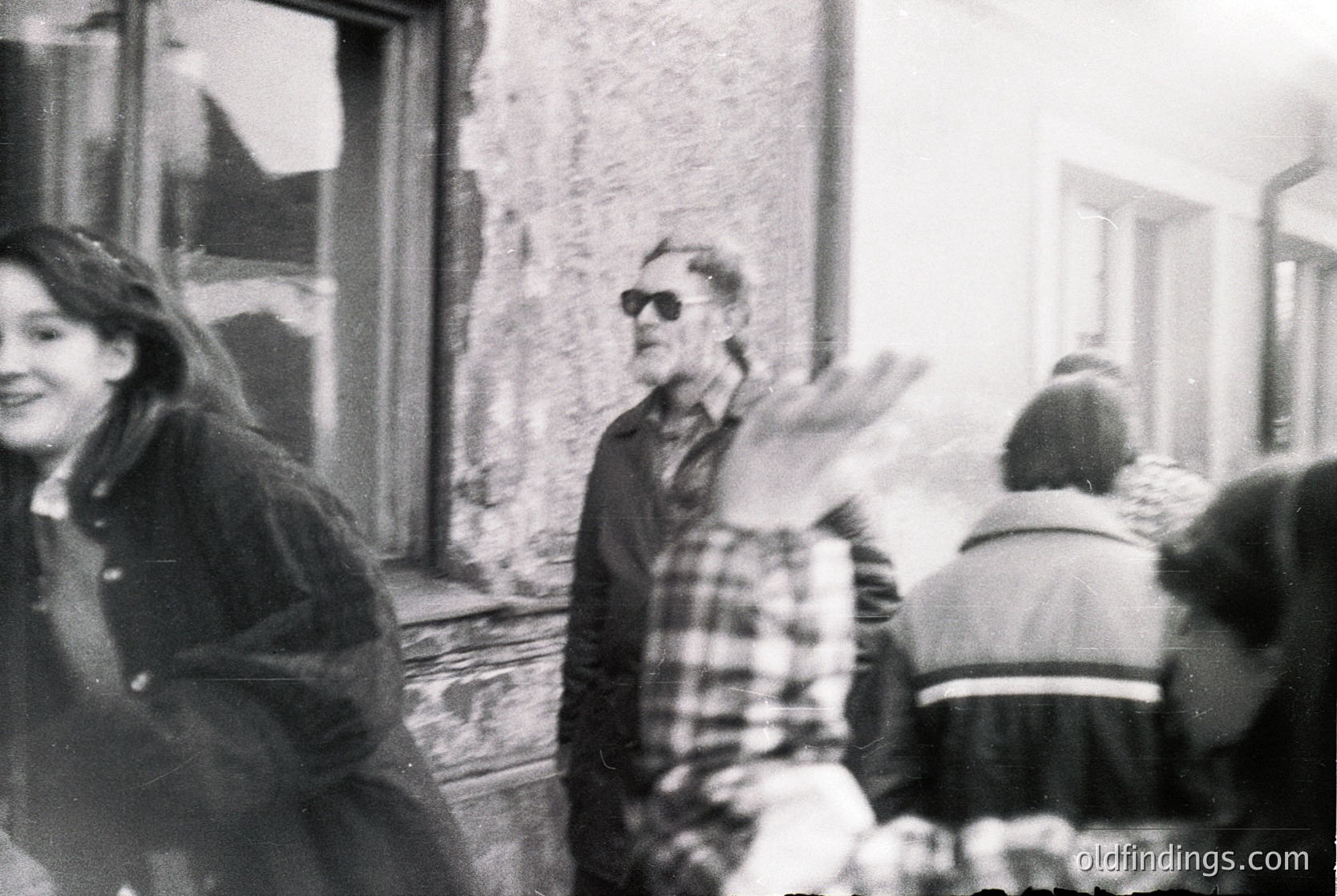 Black-and-white street scene featuring a central figure in a plaid shirt and sunglasses gesturing with arms outstretched. Surrounding individuals appear engaged in conversation or activity, with one woman smiling at the camera. Weathered urban architecture—peeling walls and closed shutters—suggests a mid-20th-century European setting. Candid, informal atmosphere captures daily life.