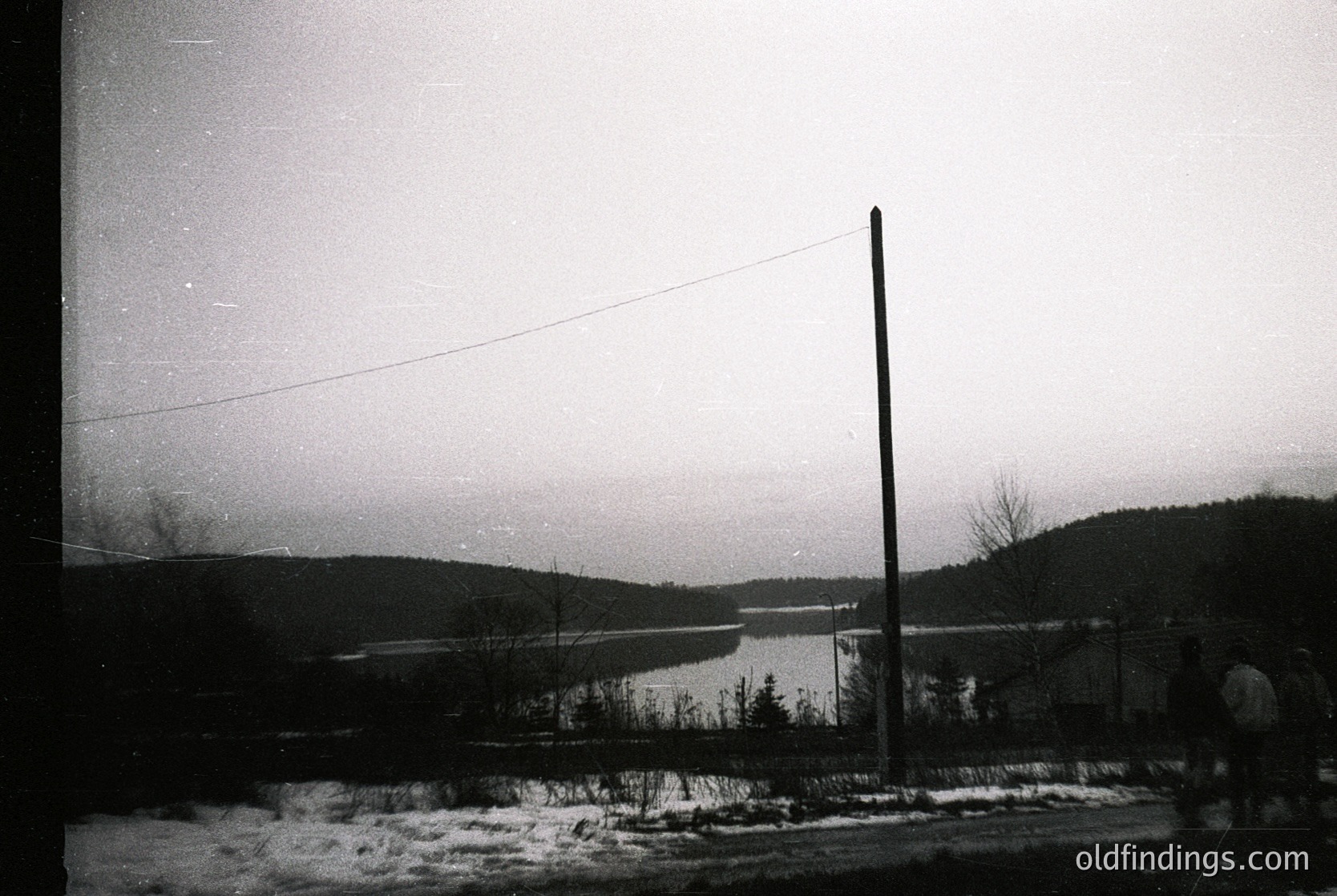 Vintage black-and-white landscape featuring a frozen lake bordered by leafless trees and rolling hills. A lone utility pole and power line dominate the right foreground, suggesting rural electrification. Snow blankets the ground, indicating winter. Likely taken in the mid-20th century.