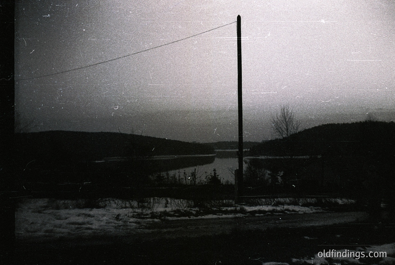 Vintage black-and-white winter landscape featuring a lone utility pole framing a frozen lake and forested horizon. Snow blankets the ground, creating a stark contrast with the dark sky. Likely mid-20th century, evoking nostalgia for rural winter scenes.
