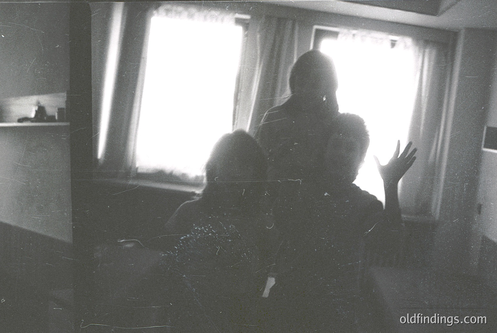 Silhouetted figures in a dimly lit interior, framed by sunlight streaming through curtains. Likely mid-20th century based on lighting and grain. Possible indoor gathering or waiting area.