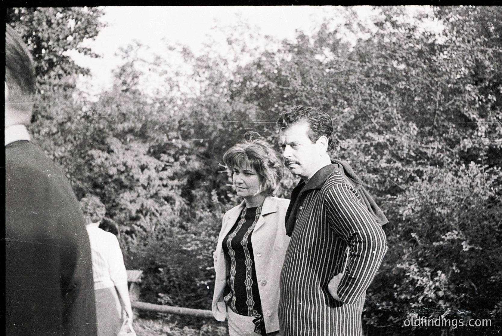 Black-and-white candid shot of three individuals in a wooded area, likely mid-20th century. Central figures wear striped blazers and a sleeveless top with geometric patterns, suggesting 1960s fashion. Dense foliage and blurred background indicate an outdoor event or gathering. Partial view of a fourth person on left adds depth.
