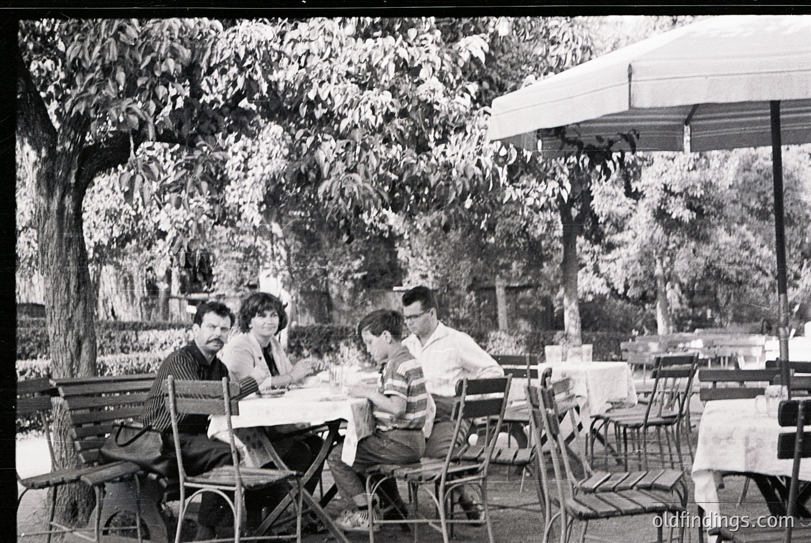 Mid-century outdoor café scene with four seated patrons at metal-framed tables under large umbrellas. Lush greenery and mature trees frame the setting, suggesting a European or Mediterranean vibe. Clothing and hairstyles suggest the – era. é