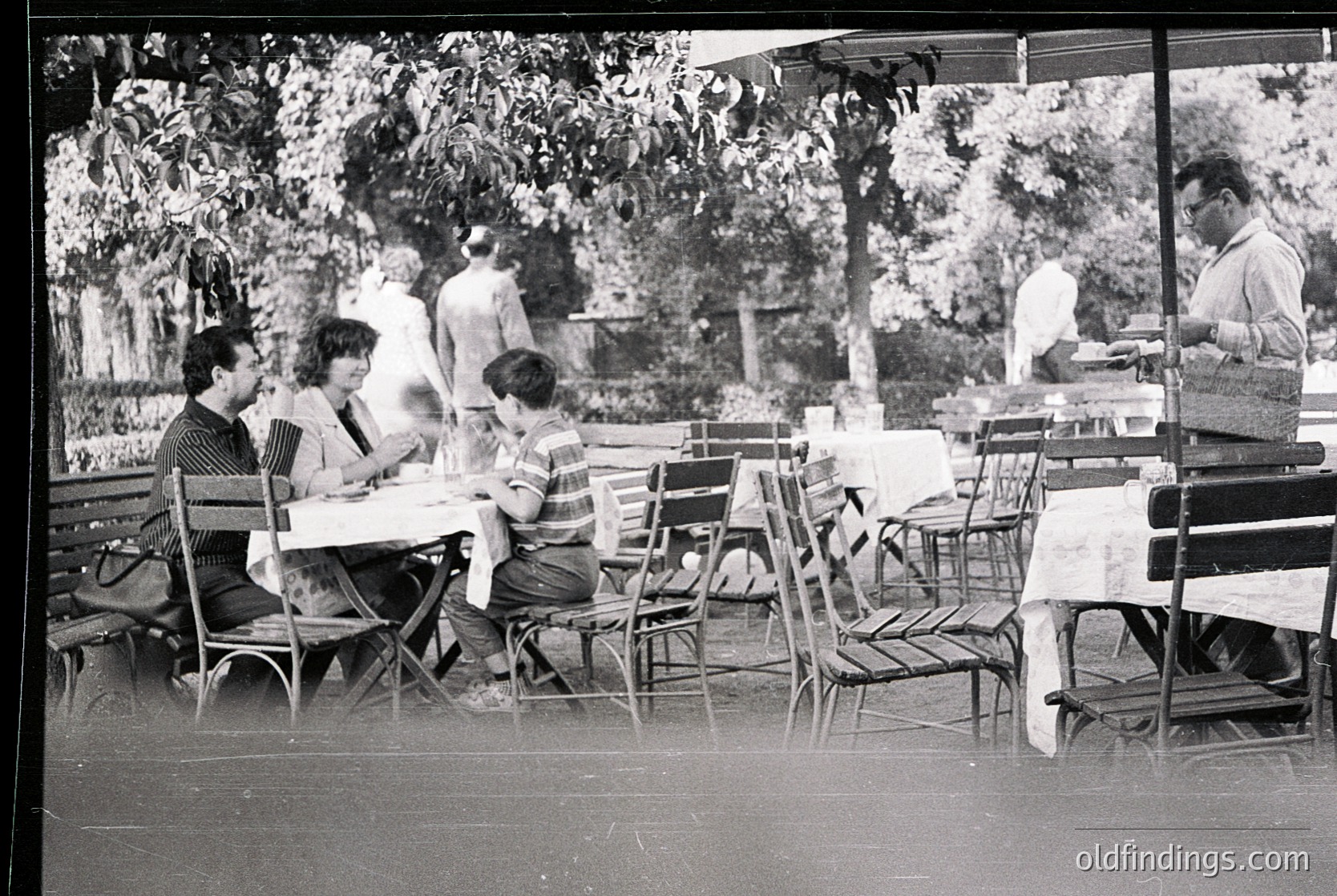 Mid-century outdoor café scene with metal-framed chairs and tables under leafy trees. Three adults and a child seated, engaged in conversation. Staff in white aprons serve food. Likely European, 1950s–1960s. é