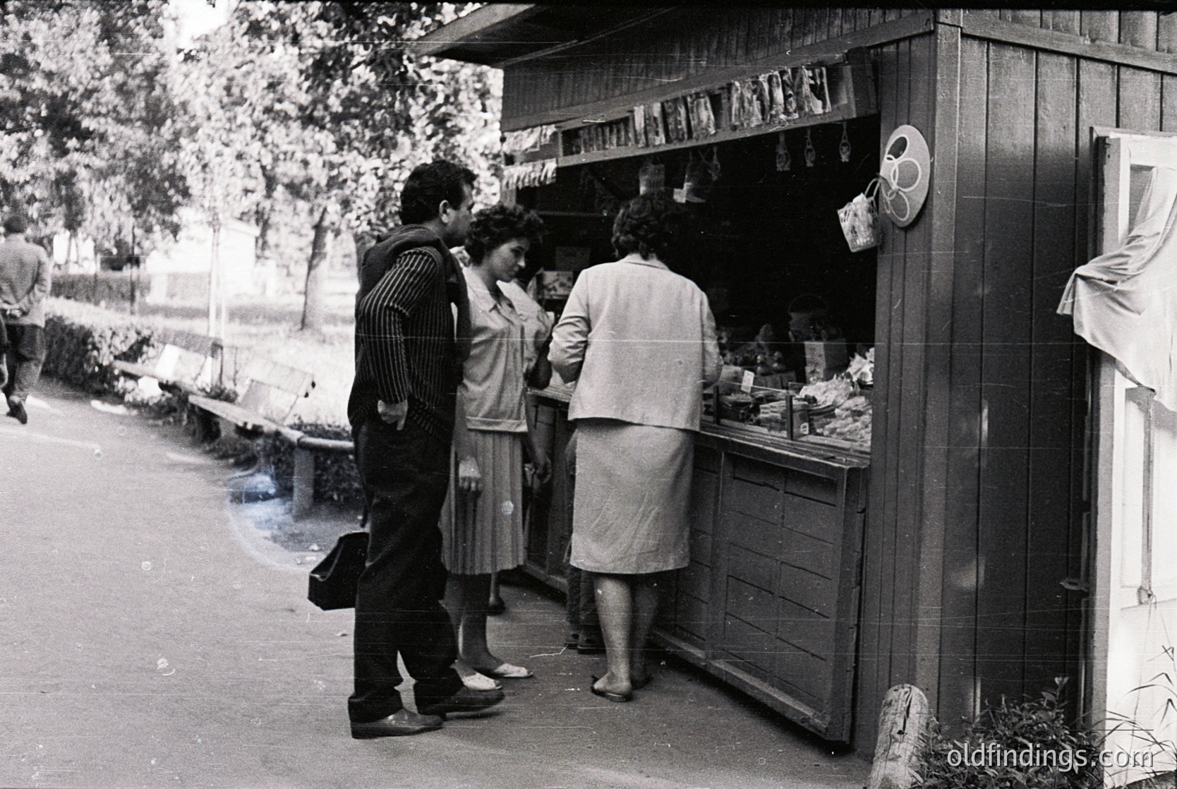 Street-side kiosk with vintage 1960s-70s design, featuring shelves stocked with jars, bottles, and packaged goods. Three people—man in striped shirt, woman in skirt, and another in a light coat—engage near the counter. Black-and-white photo captures mid-century urban life.