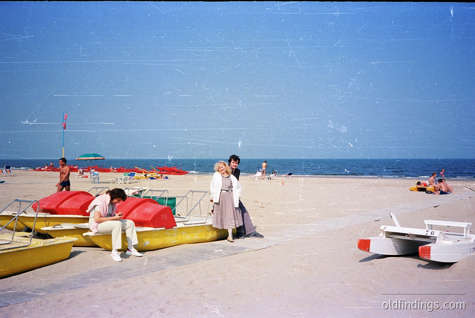 Vintage seaside scene with mid-20th century beachgoers. Yellow rowboats, red umbrellas, and a lifeguard tower frame a sandy shore under clear skies. Two women in summer dresses pose near the water’s edge, while others relax or swim. Classic beachwear and boats suggest mid-century coastal leisure.
