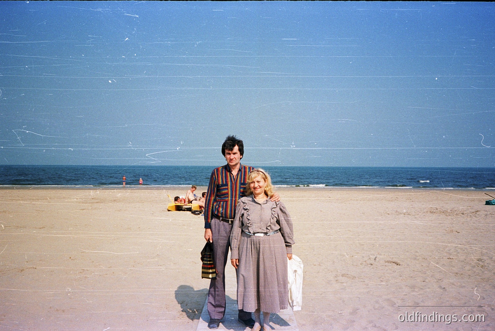 Two individuals pose on a sandy beach under clear skies, likely mid-20th century. The man wears a striped sweater and patterned pants; the woman wears a knee-length dress with a belt. A boat and distant figures suggest a seaside resort. Vintage color tones and composition evoke nostalgia.