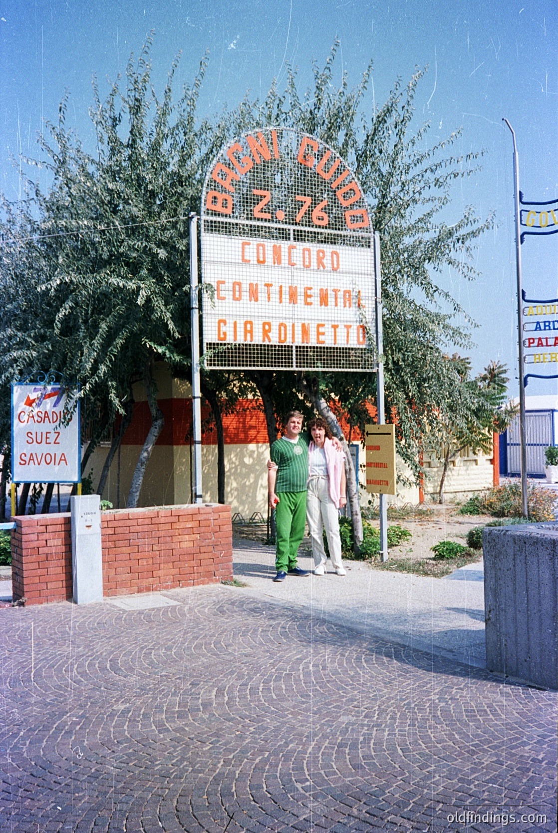 Vintage airport departure board displaying flights to Rome Ciampino (AZ 760), Concord, Continental, and Gardimetto. Two passengers in 1970s-style attire (green pants, white tops) stand near a palm tree-lined entrance. Brick planters and tiled pavement suggest mid-century modern design. Signage includes "Casadia Suez Savoia" and "Palais" in background.