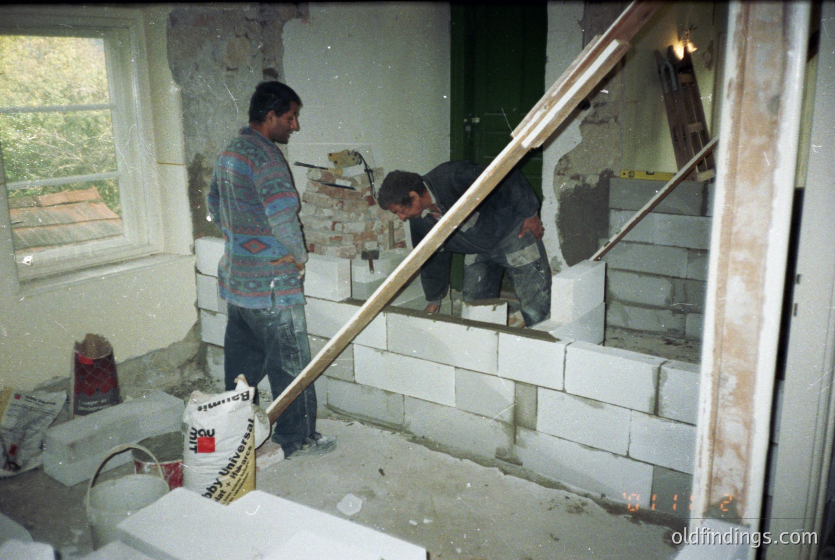 Two construction workers in a mid-renovation interior, likely residential. One stands holding a trowel, wearing a patterned shirt and jeans; the other kneels, using a level against a partially demolished wall. Exposed brickwork and debris suggest structural updates. White paint cans and tools indicate plastering or drywall work. Natural light streams through a window with a white frame.