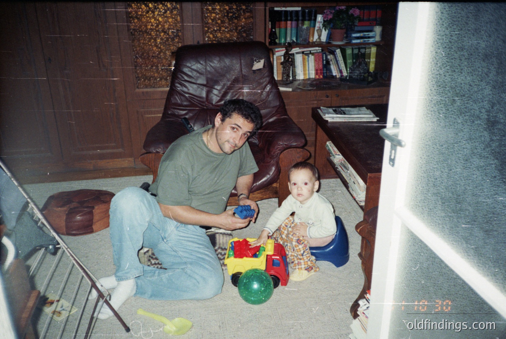A man and child play with colorful plastic toys on a carpeted floor in a mid-century interior. Wooden bookshelves filled with books flank a brown leather armchair. The scene reflects a domestic setting from the **1980s–1990s**.