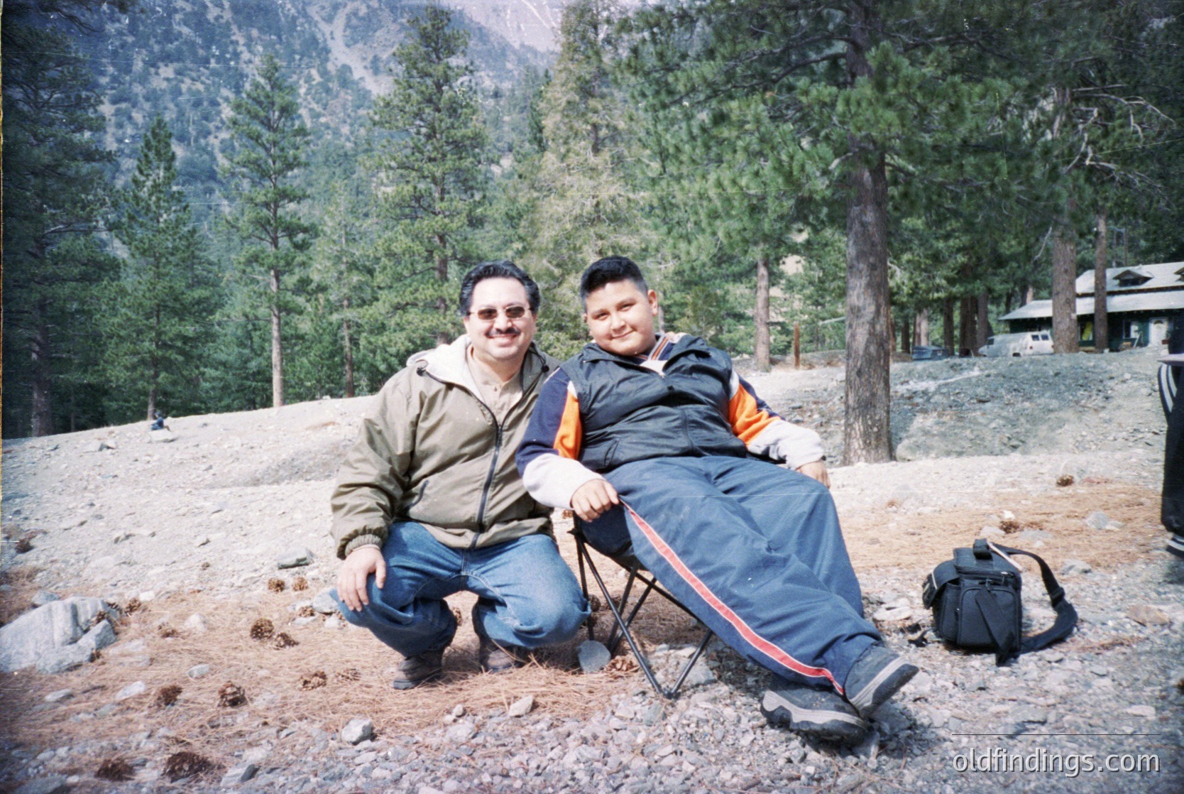 Two men pose outdoors in a forested mountainous area, likely late 1990s–early 2000s. One kneels, the other sits on a folding chair. Snow patches and pine trees frame them; a backpack rests nearby.