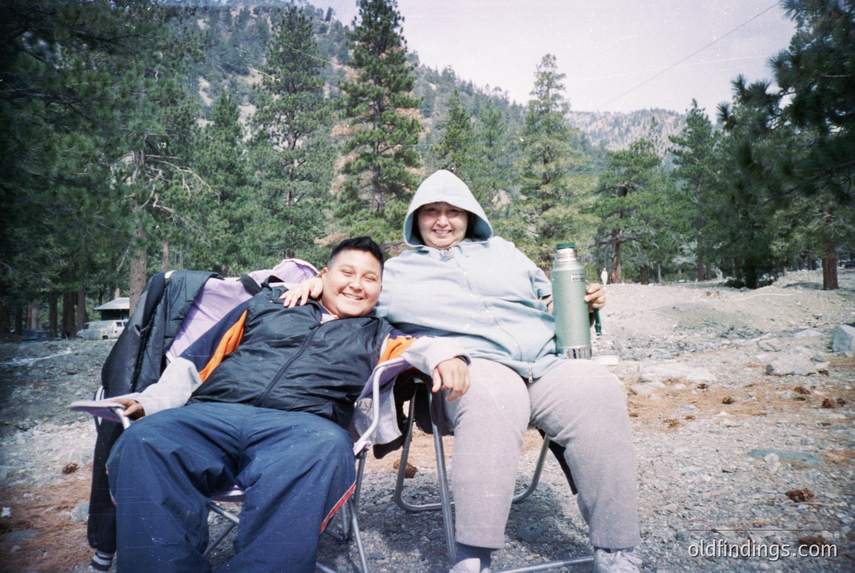 Two individuals pose outdoors in a forested area, likely during the 1990s–2000s. One wears a backpack, the other a vintage-style insulated thermos. Gravel path and pine trees frame the scene.