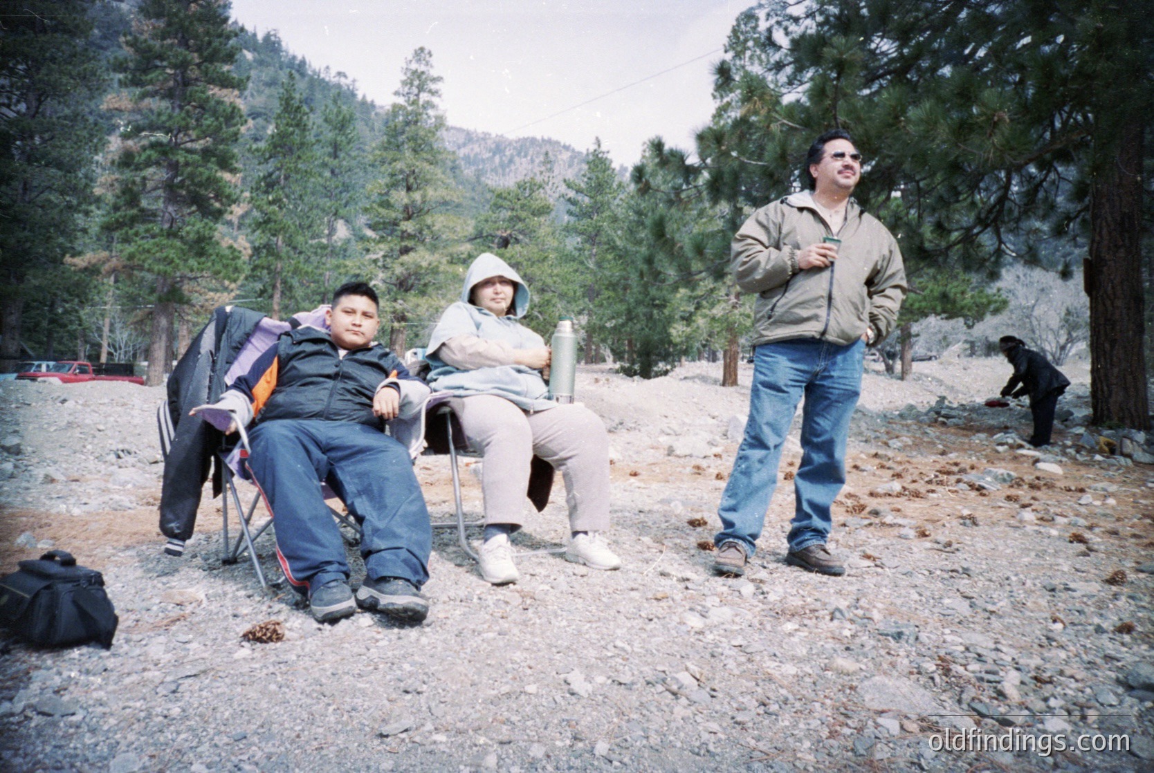 Three individuals in outdoor mountain setting, likely 2000s. Man in foreground wears beige jacket, jeans, and hiking boots; seated couple in casual outdoor wear with backpacks. Forest of coniferous trees and rocky terrain. Casual, informal group activity.