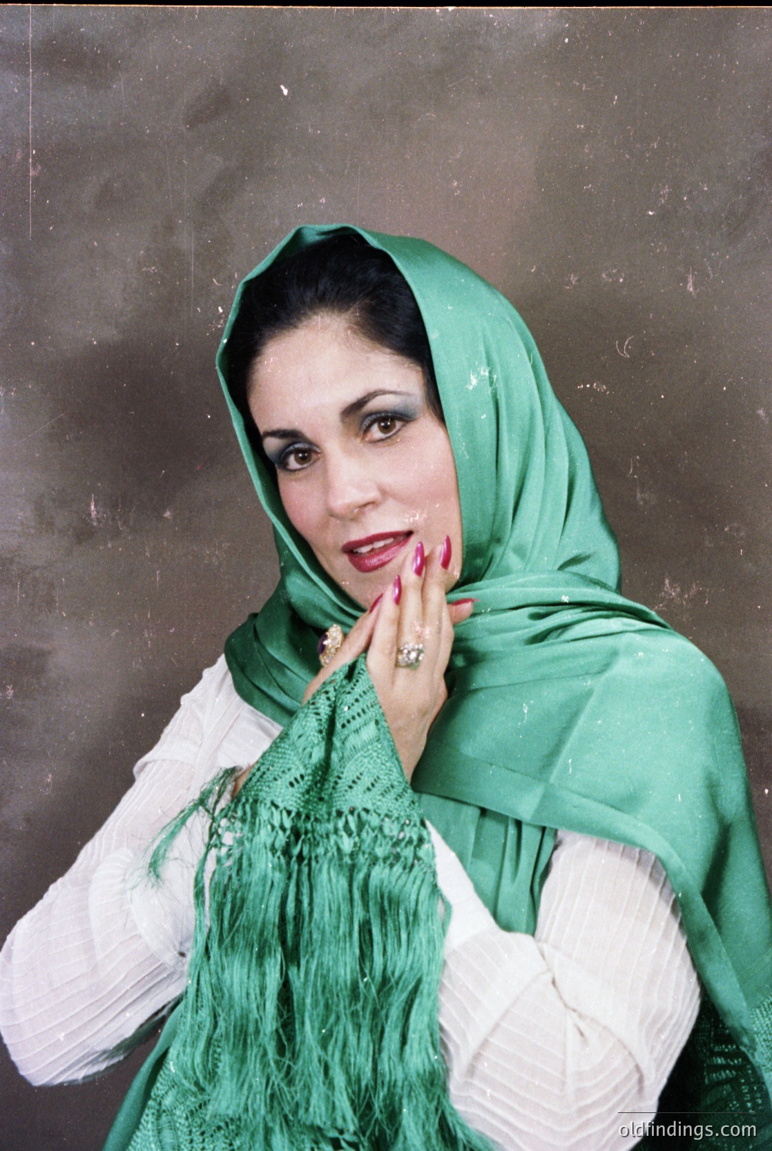 Portrait of a woman in traditional Middle Eastern attire, featuring a green headscarf and white embroidered garment. Her pose suggests a candid, intimate moment. Likely 20th-century fashion, possibly –. Background suggests an indoor setting, possibly a studio.