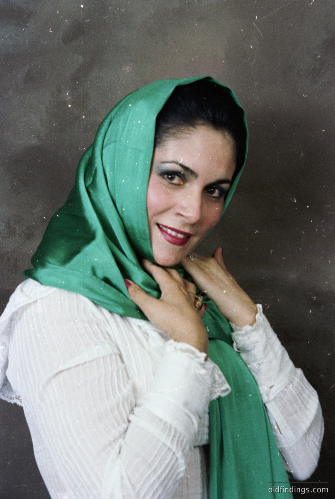 Portrait of a woman in a green headscarf and white blouse, posed with a relaxed hand gesture. The lighting suggests a studio setting with soft shadows. Style evokes mid-20th century fashion, likely 1950s–1960s.
