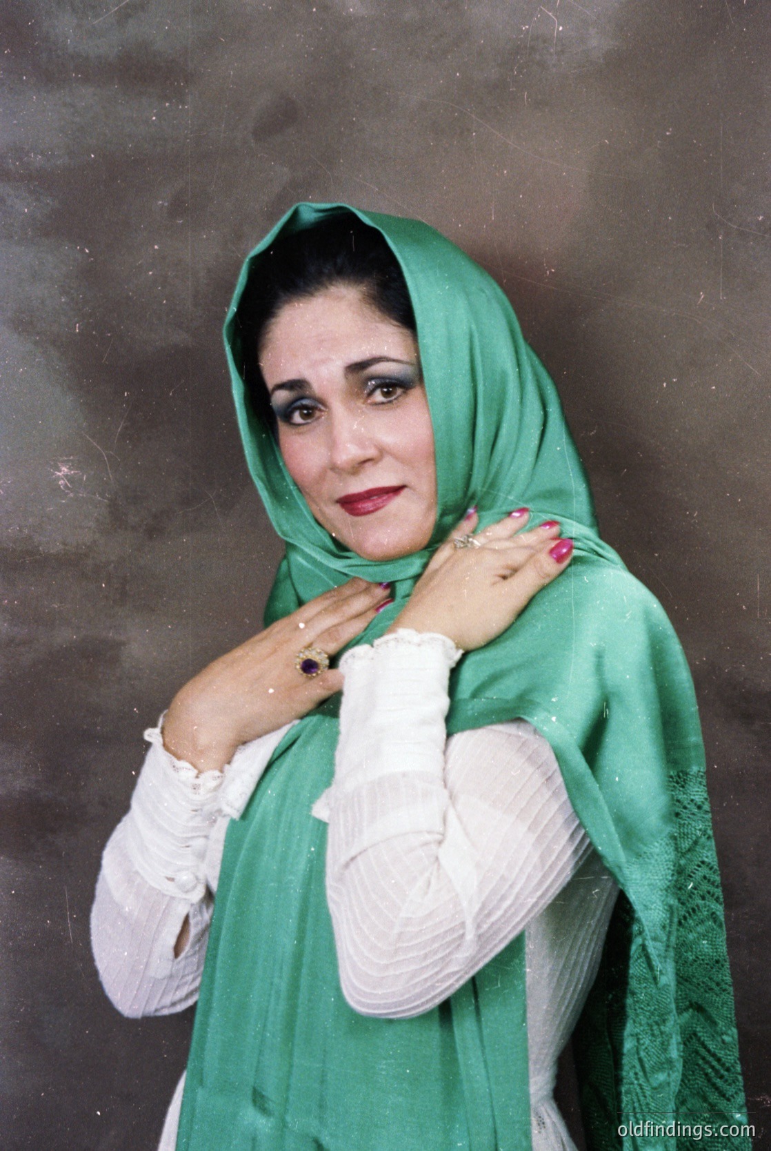 Portrait of a woman in traditional attire, likely from the Middle East or South Asia. She wears a bright green headscarf and long-sleeved white garment with intricate embroidery. Her pose suggests a formal or professional setting, possibly from the late 20th century. [Elegant woman in traditional green headscarf and embroidered white garment, striking pose with hands clasped. Likely 1980s–1990s South Asia/Middle East. ]