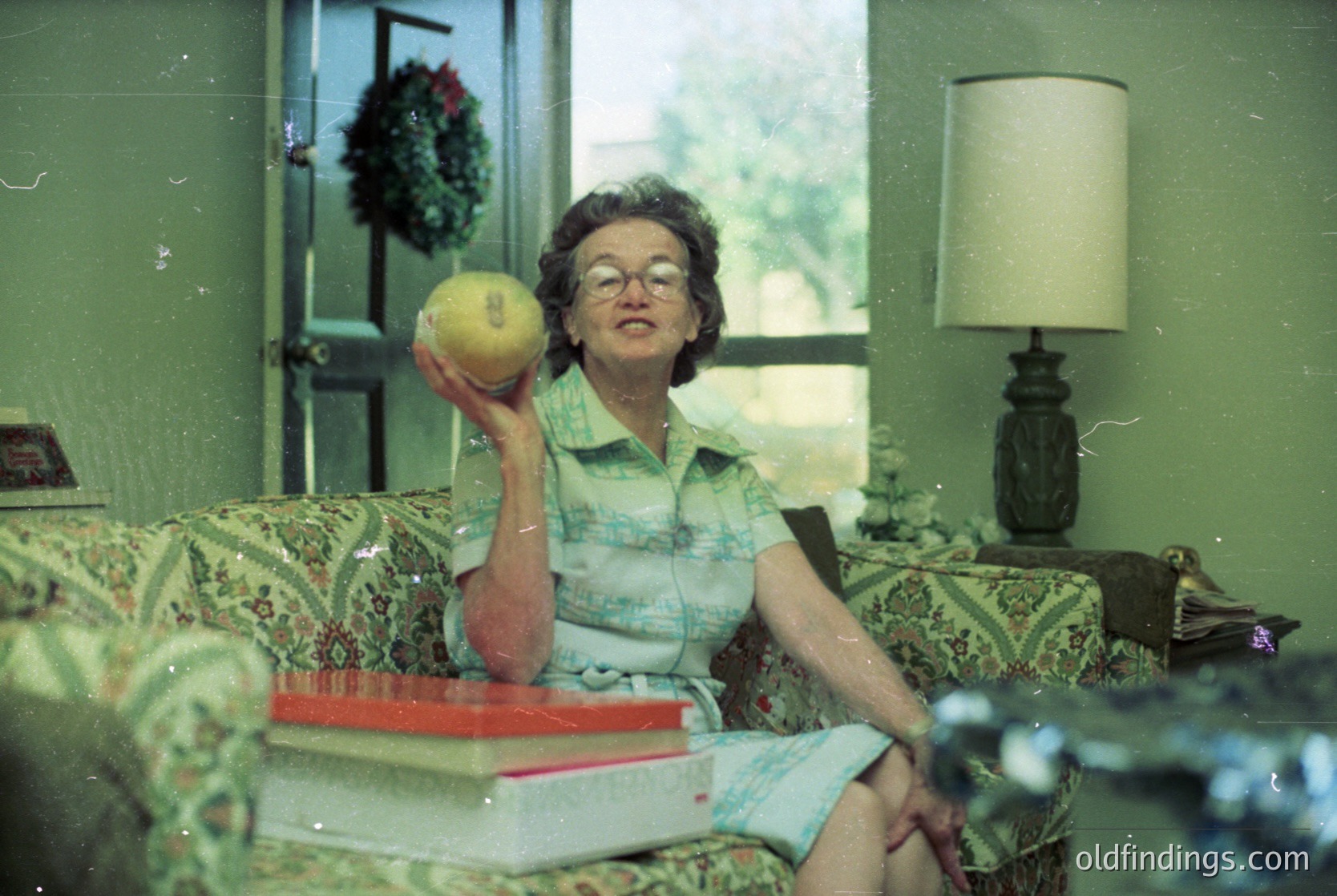 Mid-century living room interior featuring a woman in a striped blouse and light-colored skirt, holding a tennis ball. Floral-patterned sofa with a stack of books (red cover) beside her. Vintage lamp with dark base and white shade on a side table. Wreath on a door frame and large window revealing greenery. Likely 1960s–1970s domestic setting.
