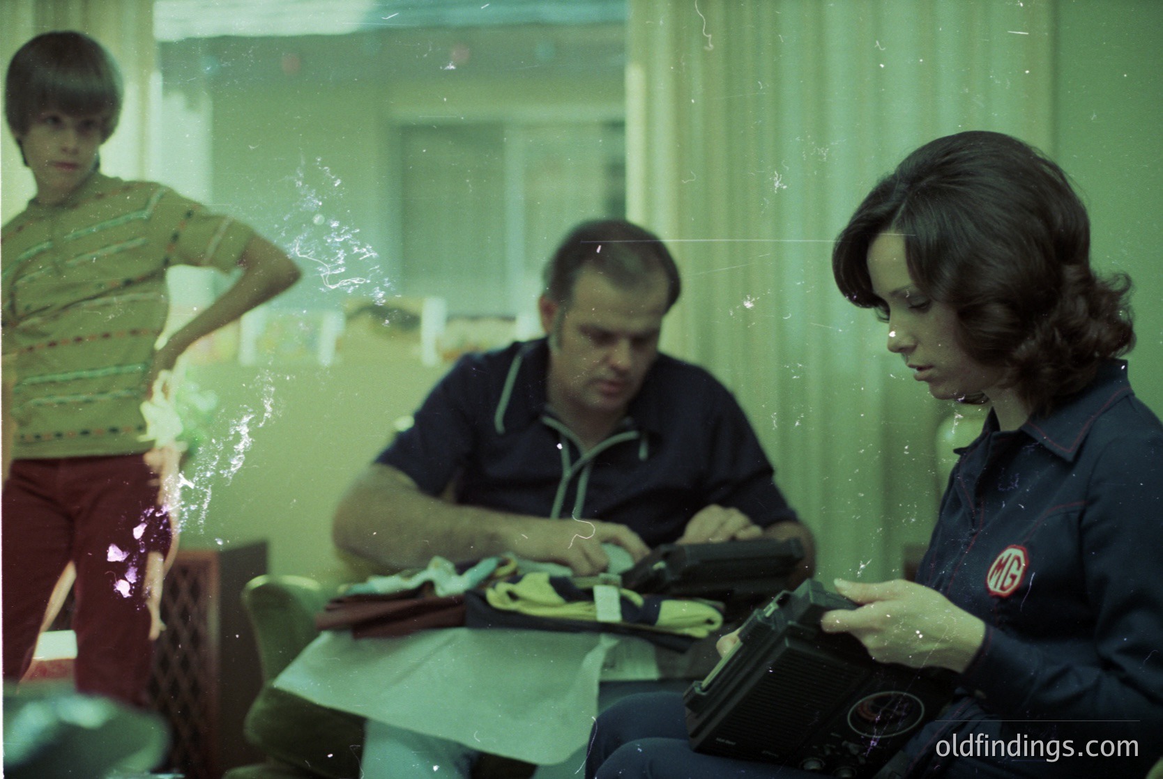 Vintage indoor scene featuring three individuals seated at a table with a typewriter. Man in striped shirt and woman in dark uniform (likely a 1970s hotel or office setting) appear to be working. Boy in green shirt stands behind them. Warm lighting and patterned curtains suggest a mid-century European workspace.