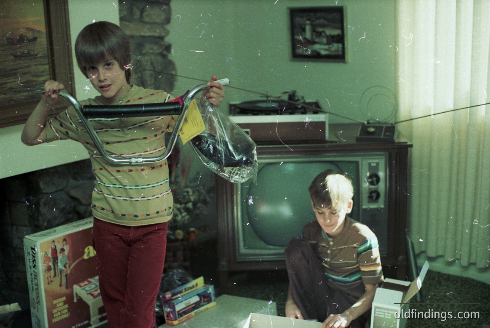 Vintage indoor scene featuring two boys in 1970s-style striped sweaters and red pants. One boy holds a transparent plastic bag, while the other sits on a floor with an open box. A CRT TV and framed maritime artwork fill the background, suggesting a mid-century living room. Decor includes a shelf with figurines and a boxed toy.