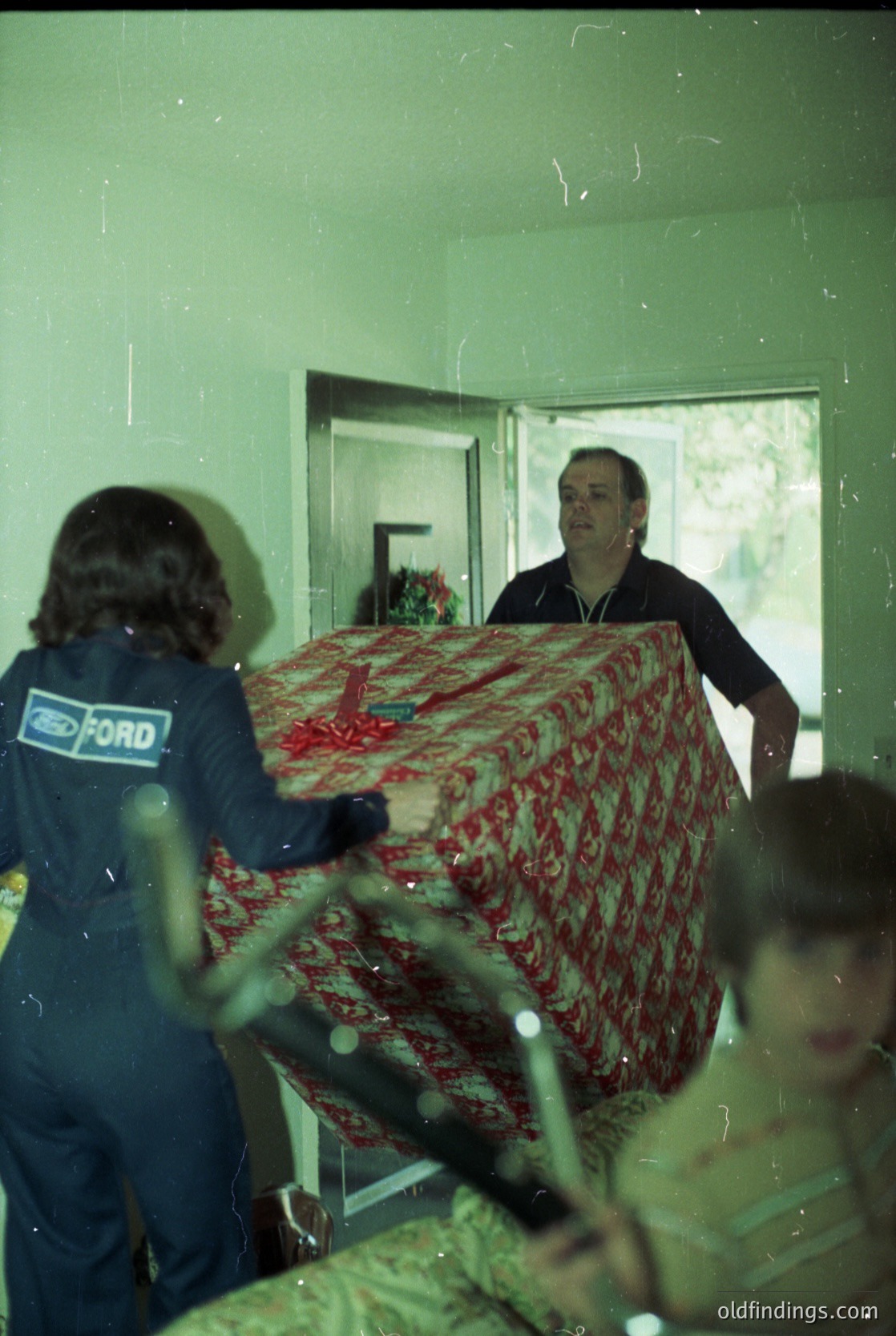 Vintage indoor scene featuring two men handling a large, patterned red-and-white fabric (likely a bedspread or curtain) in a residential setting. One man wears a Ford-branded uniform, suggesting automotive industry work. The other wears a dark suit. Blurred figures in the background indicate a busy household. Likely 1960s–1970s, Western industrialized country.
