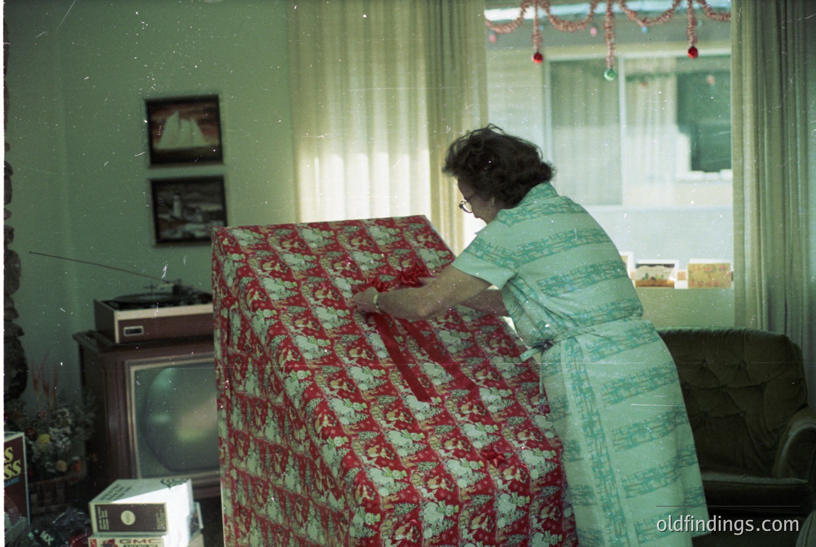 Mid-century living room interior featuring a woman in a light-colored dress dusting a floral-patterned sofa (red/cream). Black-and-white radio and vintage TV on left. Curtains with red tassel garland above. Likely 1960s–1970s domestic setting.
