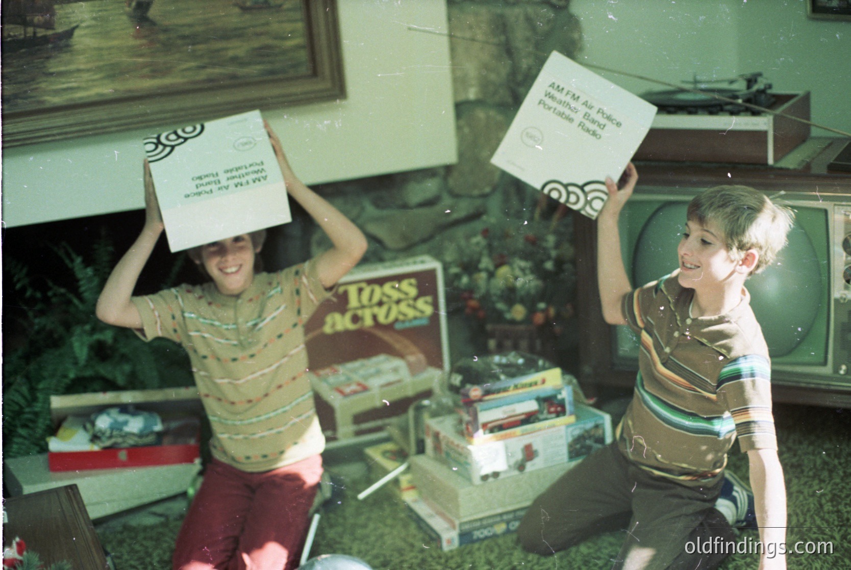 Two boys in 1970s-style striped shirts balance cardboard boxes on their heads indoors, likely a 1970s living room. Boxes labeled "AM/FM Portable Radio" and "Toss 'n' Actus" suggest early gaming/entertainment trends. Wooden shelves, a TV, and framed art hint at mid-century home decor.
