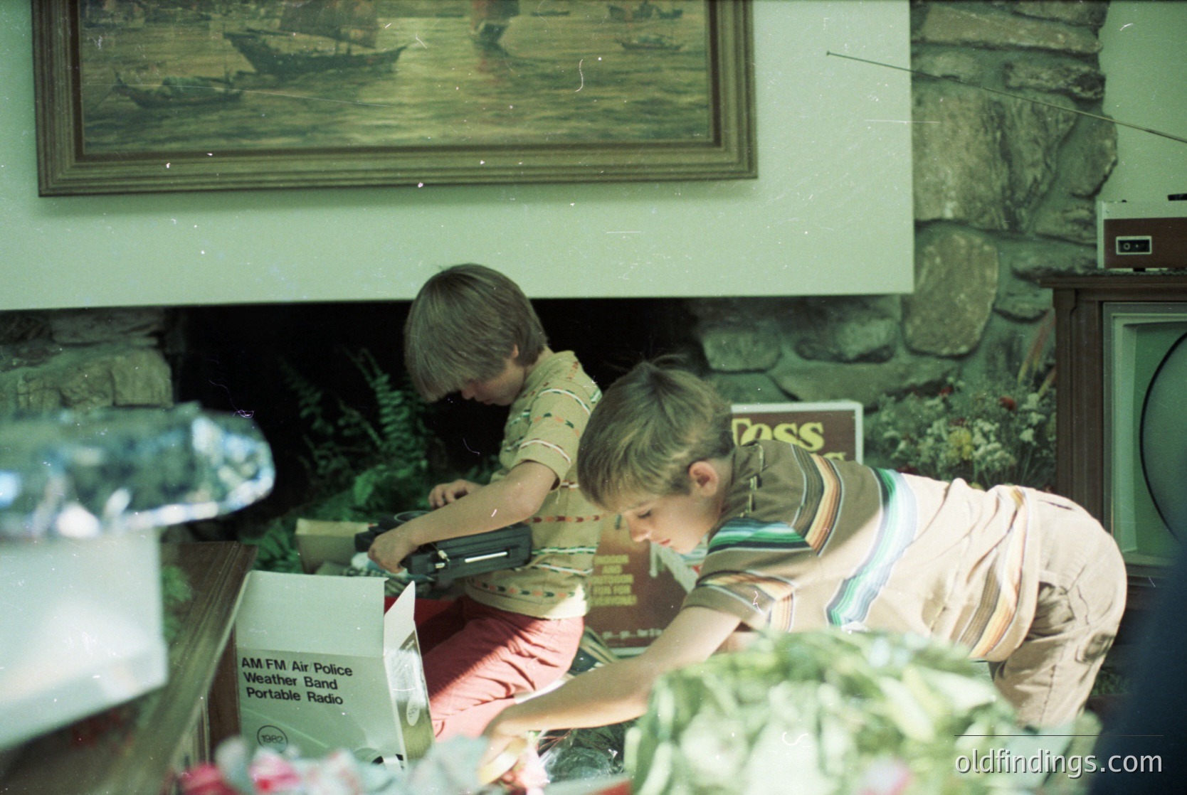 Vintage indoor scene featuring two children engrossed in crafting near a stone fireplace. One child wears a striped shirt, the other a floral top. A framed maritime painting and a "PASS" sign adorn the wall. A portable radio box (AM/FM Police Weather Band) rests on the table. Floral arrangement and vintage decor suggest a mid-20th-century home setting.