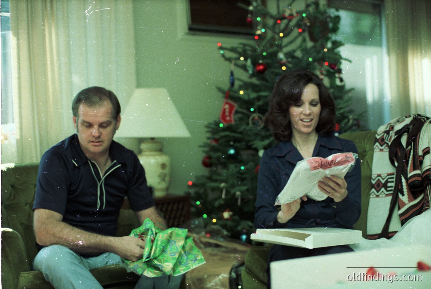 Vintage 1970s holiday scene: man in loose-fitting sweater and jeans unwraps gift beside woman in wide-collar blouse holding wrapped package. Decorated Christmas tree with red/orange ornaments and string lights fills background. Warm, intimate living room with patterned throw blanket and classic lamp.