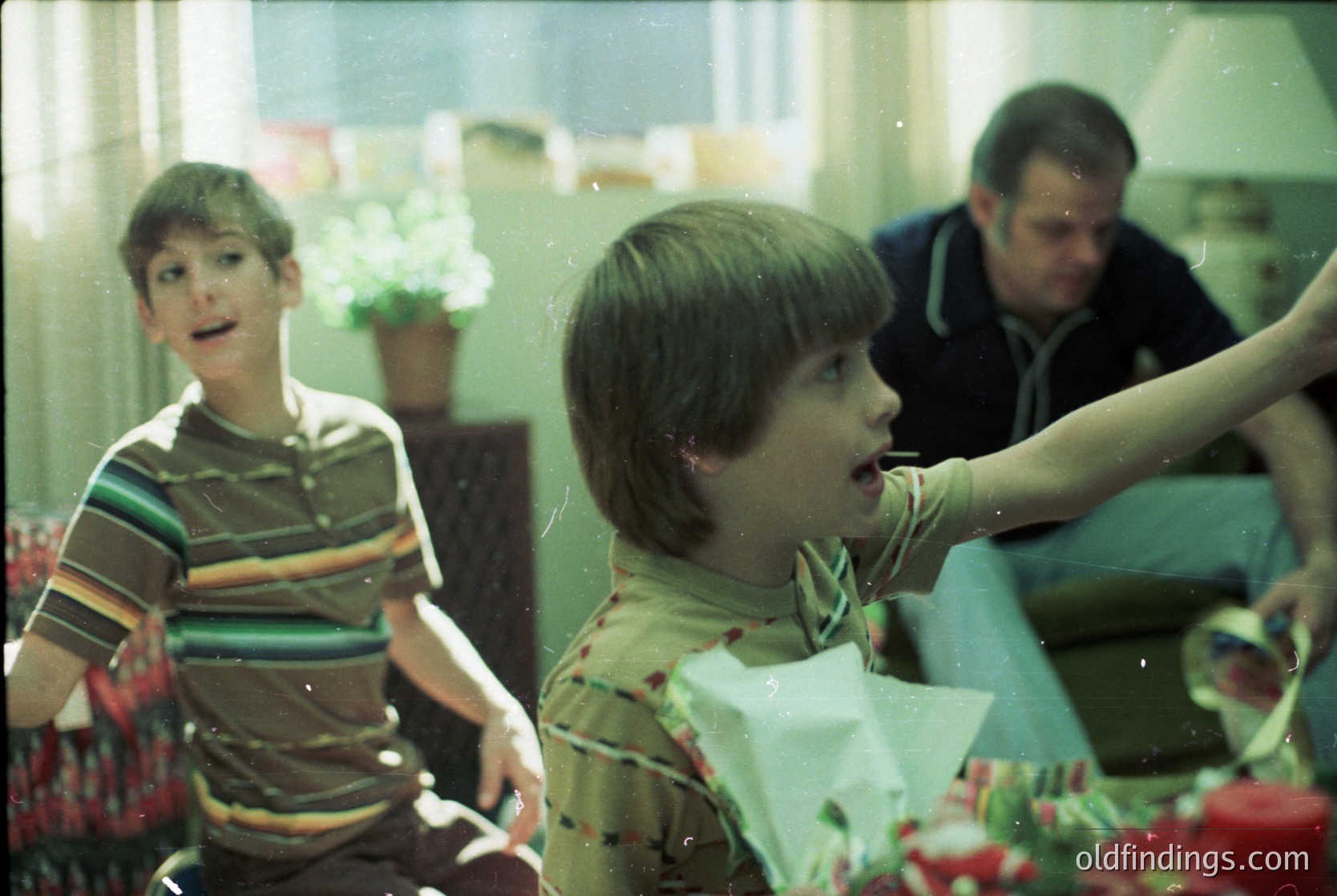 Vintage indoor scene featuring two boys and an adult in a festive setting, likely a 1970s-80s family gathering. Boys in striped and floral shirts, one holding a wrapped gift. Adult in a striped sweater, pointing toward a table adorned with red flowers. Warm, nostalgic lighting and blurred motion suggest candid, joyful moments. Ideal for historical research or vintage-themed content.