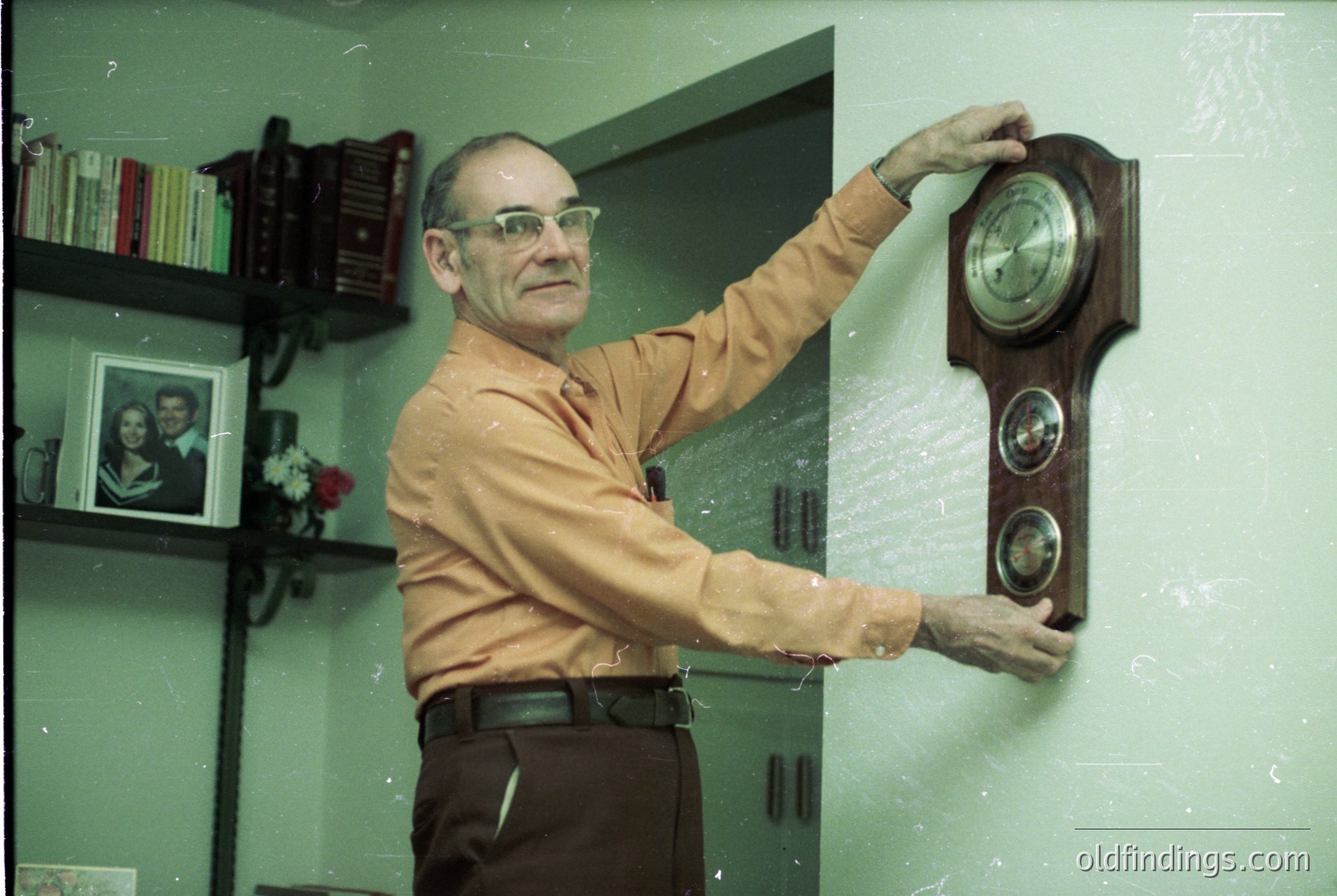 Mid-century office setting featuring a man in a light-colored button-down shirt adjusting a vintage wooden wall clock with dual dials. Shelves display framed photos, books, and a floral arrangement. Light green walls and a partial view of a desk with a typewriter suggest a 1960s–1970s professional environment.