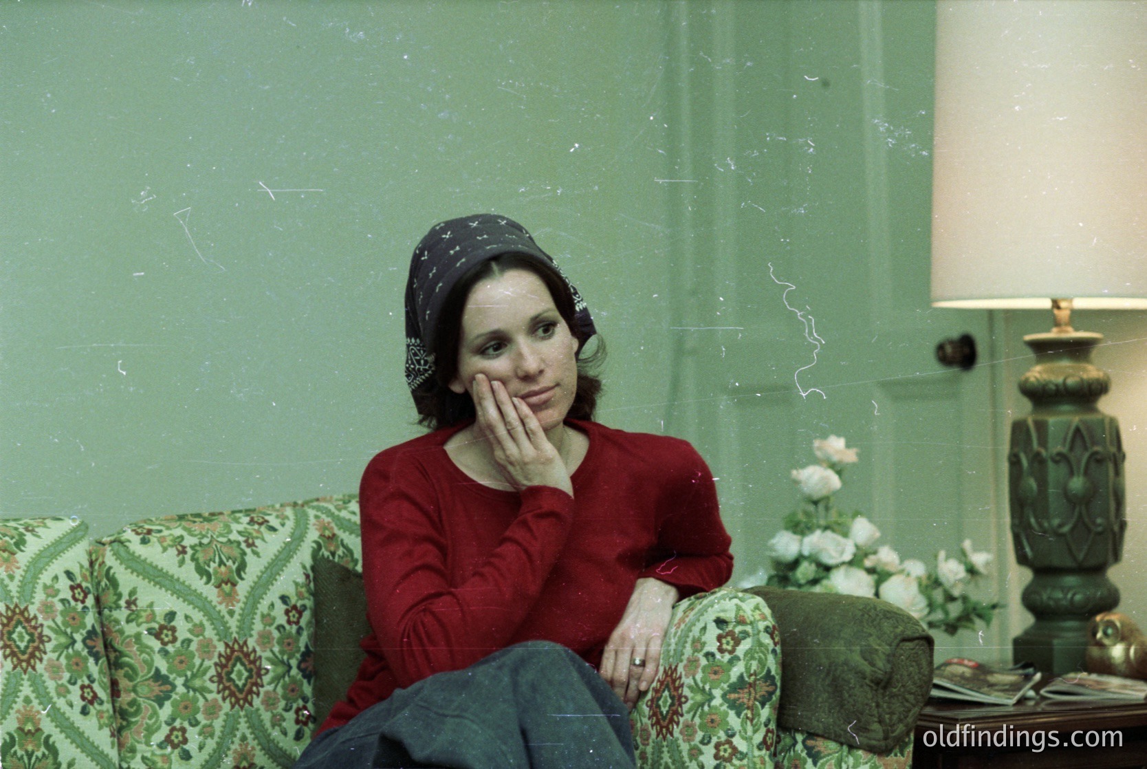 Vintage portrait of a woman in contemplative pose, seated on a floral-patterned armchair against a faded mint-green wall. She wears a red sweater, patterned headscarf, and dark jeans. A small table beside her holds a vase with white flowers and an ornate lamp. Mid-century interior design with warm lighting. Likely
