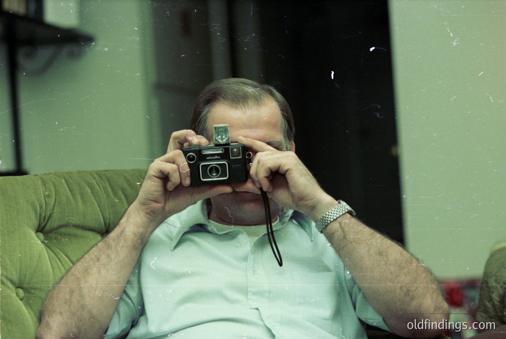 Mid-century man holds a vintage **Zeiss Ikon Contax S** camera, framed by a green armchair and muted indoor lighting. Likely 1960s–1970s, reflecting analog photography’s golden era.