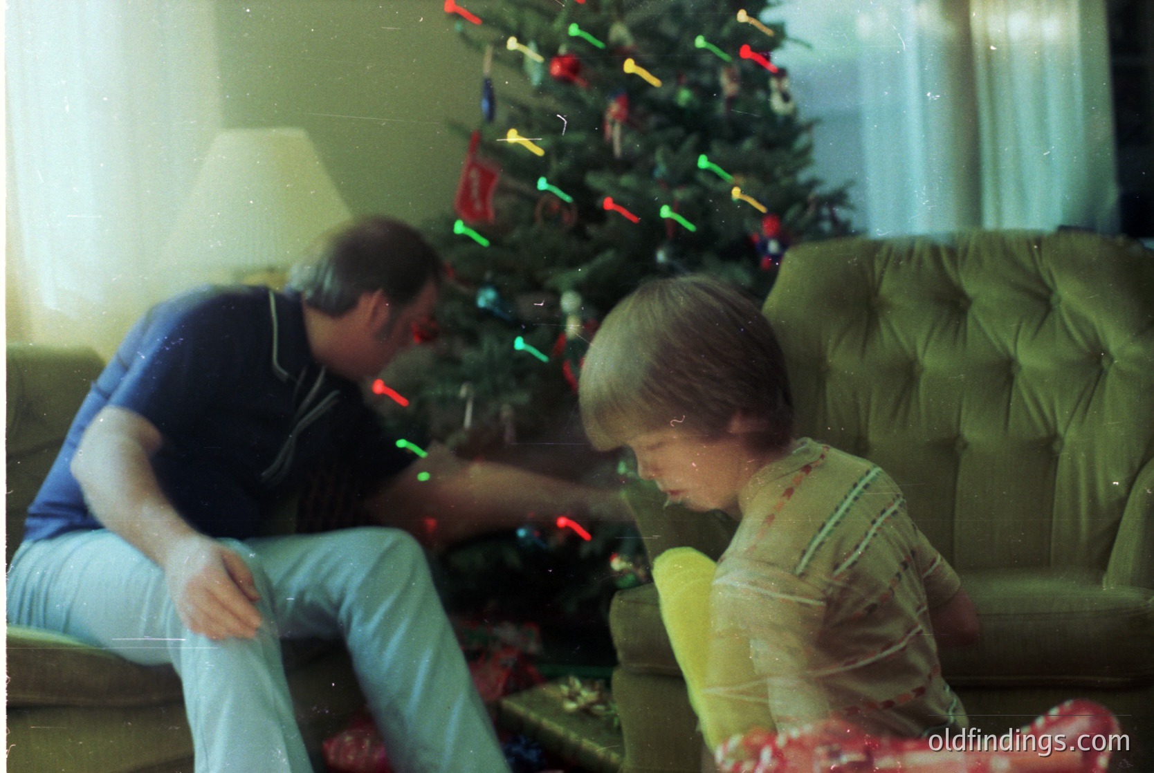 Vintage indoor scene: man and child examining wrapped gifts under a decorated Christmas tree with multicolored lights. 1970s-80s holiday decor, including a red gift bag. Warm, nostalgic family moment.
