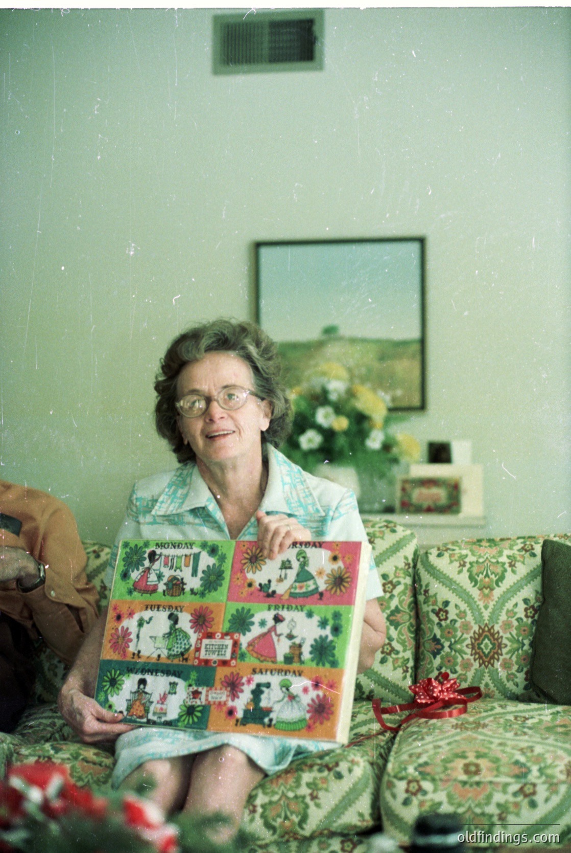 Vintage quilt featuring folk-art designs with scenes of rural life, held by an elderly woman in a mid-century living room. Floral-patterned sofa and framed landscape artwork in background suggest 1960s–1970s domestic setting.