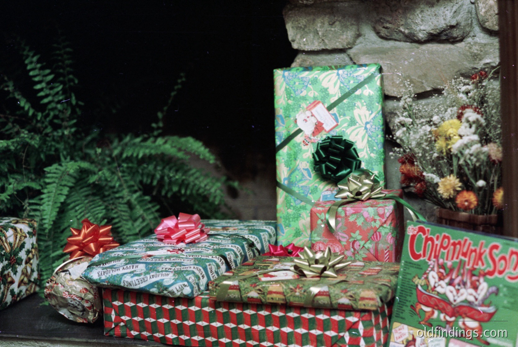 Vintage holiday scene featuring wrapped gifts with retro patterns and labels. Boxes include "Chimpmyk Soda" and festive bows on a stone ledge. Indoor/outdoor setting with greenery and dried flowers. Likely late 20th century (1970s–1980s).