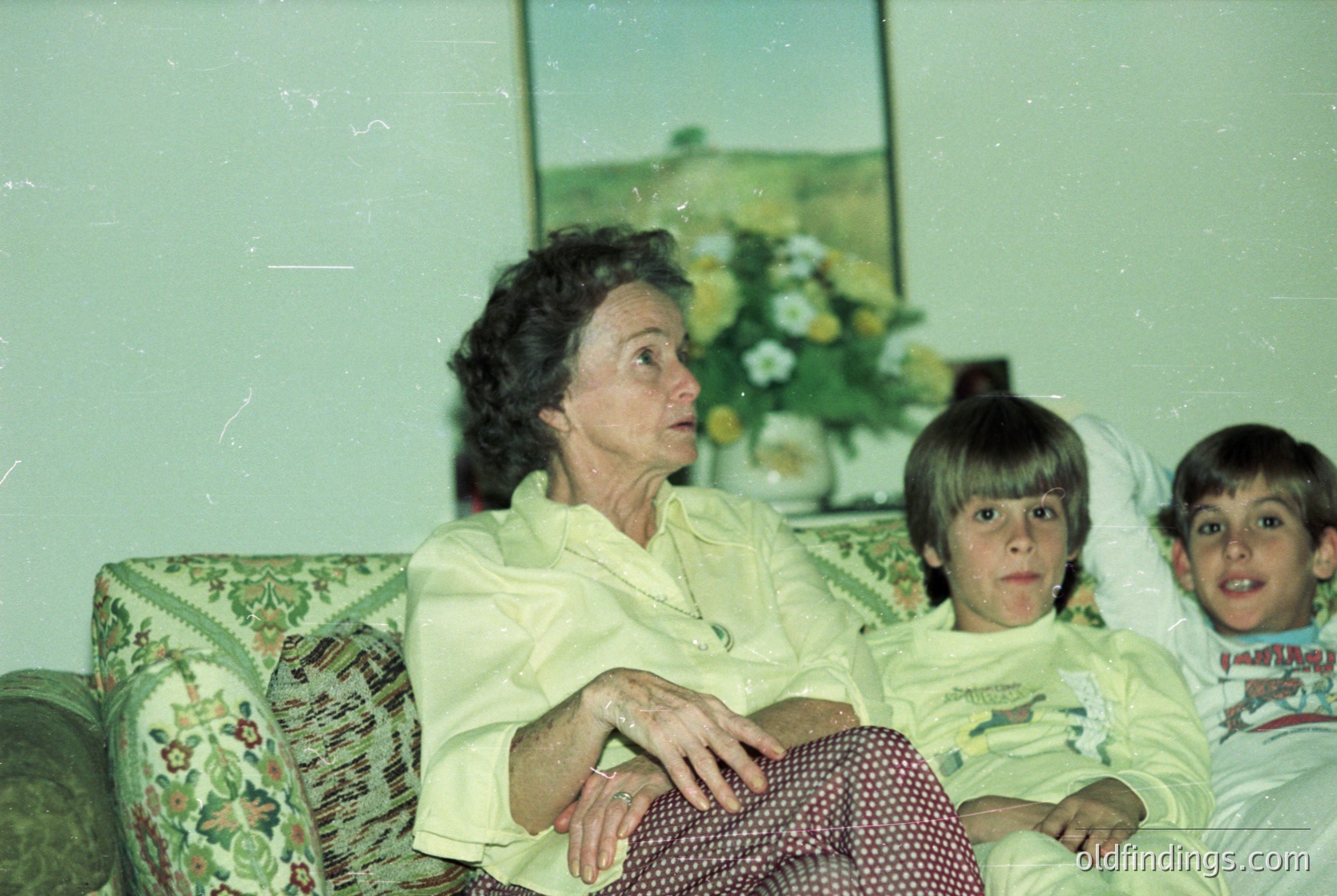 Vintage indoor portrait featuring an elderly woman and two young boys on a floral-patterned sofa. The woman wears a light-colored blouse, while the boys sport matching white T-shirts with dark designs. A floral arrangement in a vase sits on the table behind them. Likely late 20th century, mid-Atlantic region.