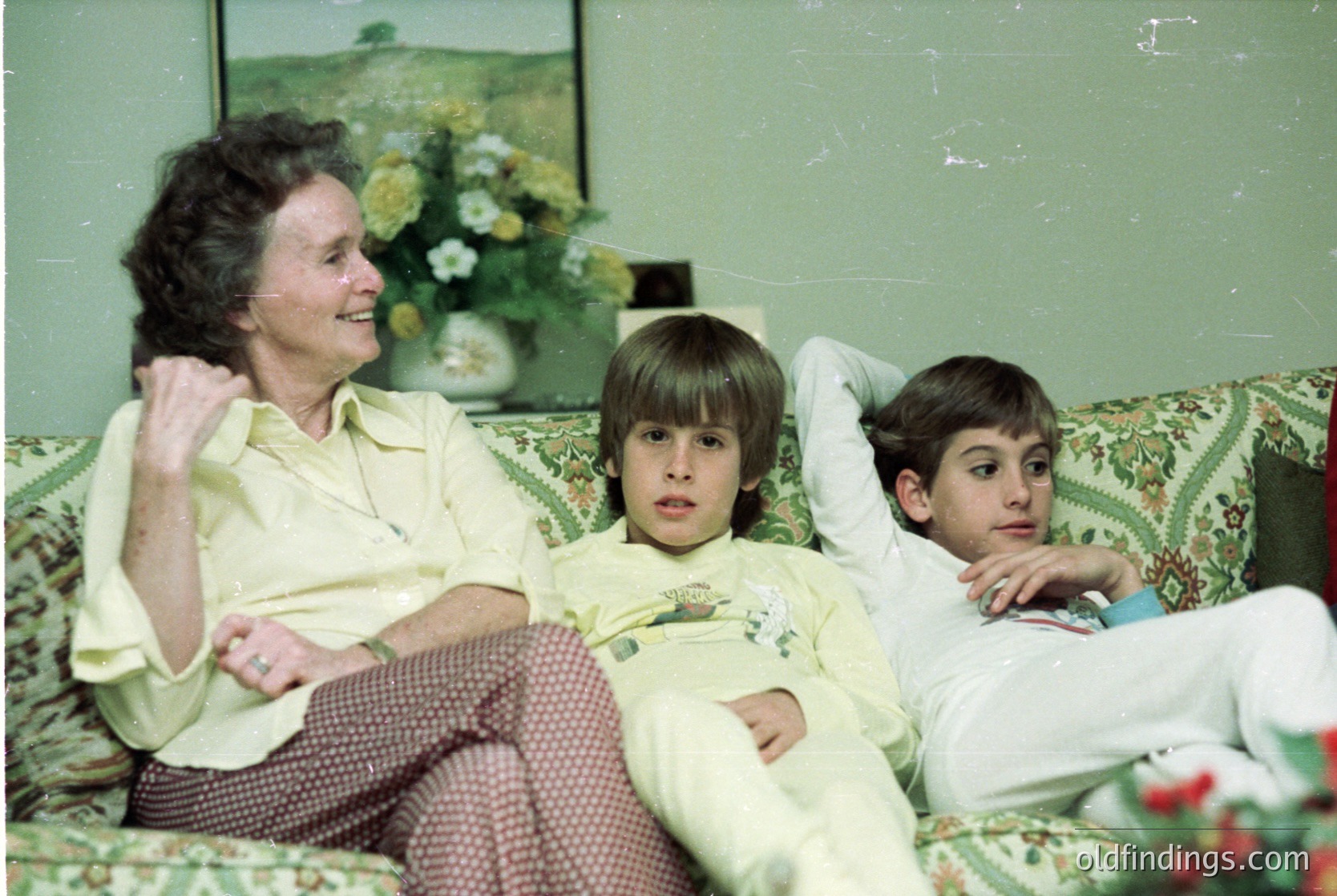 Vintage indoor portrait of a woman and two boys on a floral-patterned sofa, mid-1970s. The woman wears a light-colored blouse and patterned pants; the boys in matching short-sleeve shirts. A floral arrangement and landscape painting adorn the wall. Warm, nostalgic family moment.