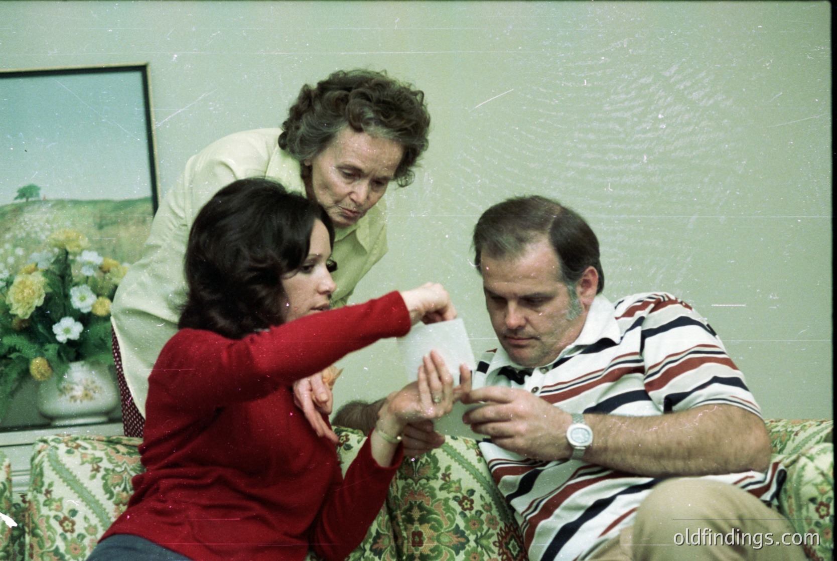 Vintage indoor scene: three adults in mid-1970s clothing (women in long-sleeve blouses, men in striped shirt) gathered around a man holding a rolled document. Woman in red assists, another woman in beige stands observing. Floral-patterned sofa and framed floral artwork on wall suggest a domestic or office setting. Warm sepia tone indicates archival or scanned photo.