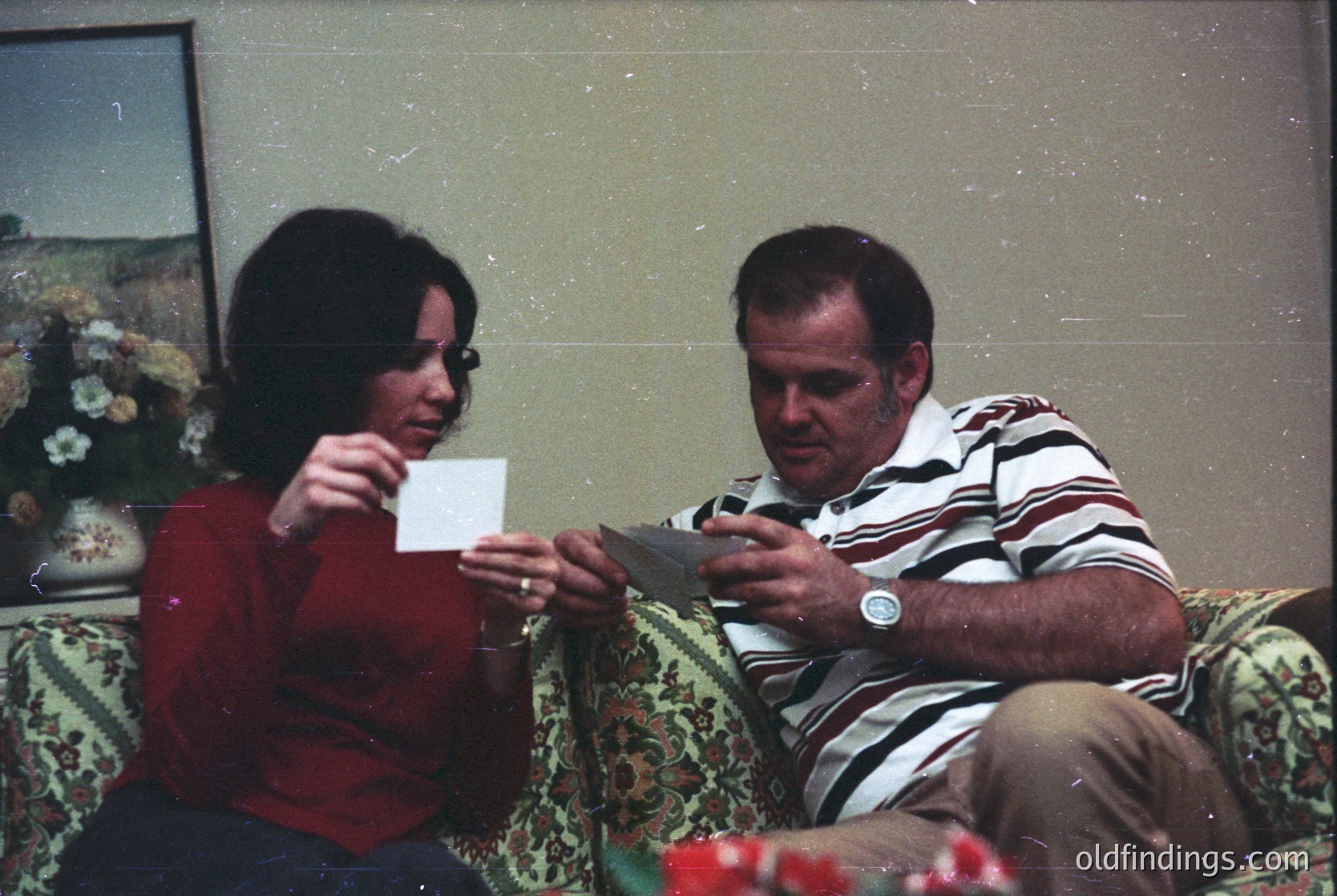 Vintage indoor portrait of two people playing cards on a floral-patterned sofa, 1970s-80s. Woman in red sweater holds a white card; man in striped shirt examines a black card. Painted landscape on wall suggests domestic setting.