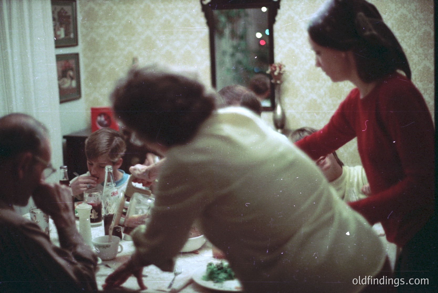 Vintage indoor gathering, likely 1960s–1970s, featuring a family dining scene. A woman in a red sweater serves food at a long table with mismatched plates, glasses, and a bottle of *Bulgaria’s* *Burgasko* beer. Framed portraits and a mirror adorn the walls, reflecting warm, lived-in lighting.