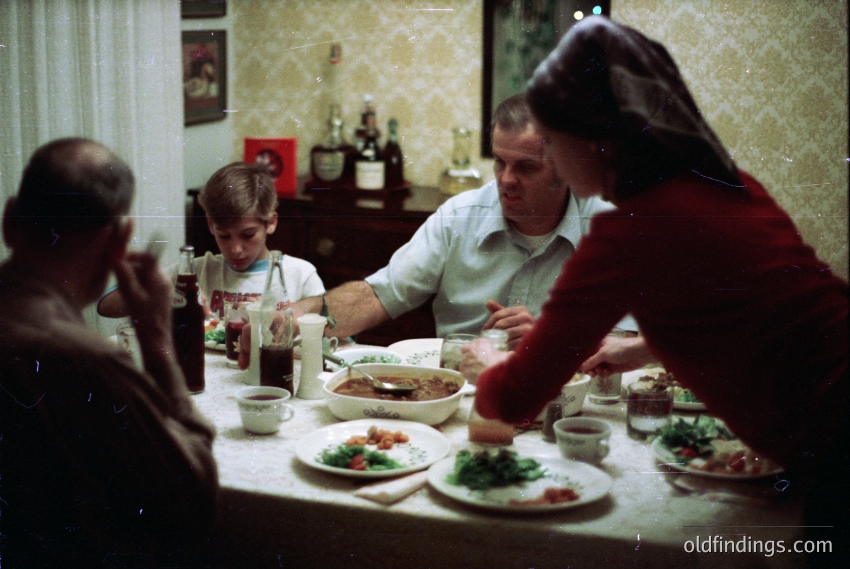 Vintage family meal scene in a mid-century home, featuring four adults and a child seated around a long table. Visible dishes include a large roasted meat platter, salads, and bread. Bottles of wine and a soda bottle suggest a casual gathering. Warm lighting and patterned wallpaper evoke 1960s–1970s domestic life.
