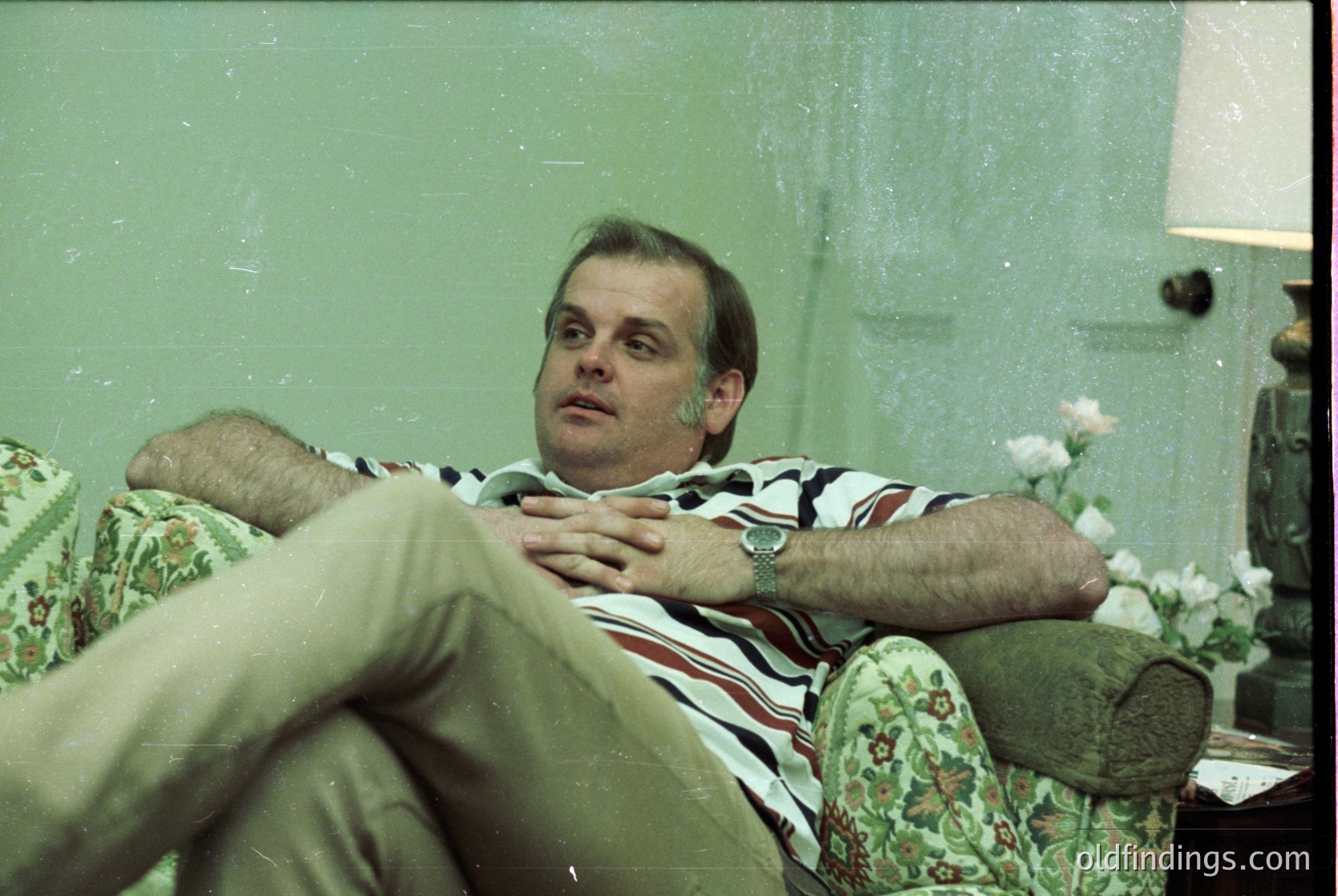 Vintage indoor portrait of a man reclining on a floral-patterned armchair, wearing a striped polo shirt and khaki pants. Soft lighting and a green-tinted filter suggest 1970s–1980s photography. Side table holds a lamp and floral arrangement.