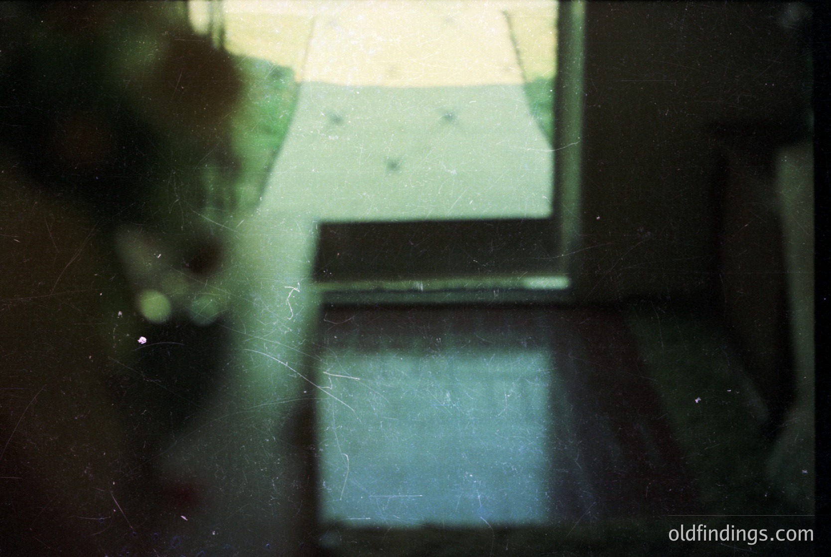 Vintage film grain captures a blurred interior through a window, revealing a dark wooden table and faint reflections of a light-colored room. Possible 1970s–1980s domestic setting, likely European. écor