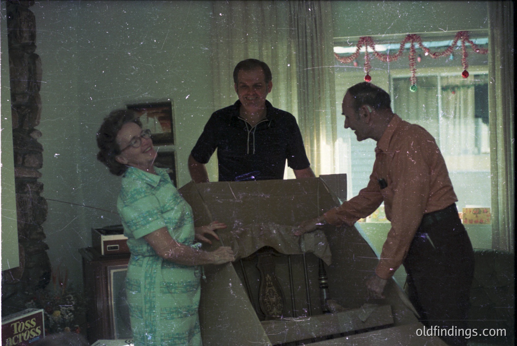 Vintage indoor scene featuring three adults decorating a Christmas tree. Woman in a floral blouse and glasses places an ornament; man in a brown shirt and tie secures it. Decor includes red garlands and a "LOSS CLOSS" sign. Mid-century home decor with wooden furniture and framed art. Likely 1960s-1970s holiday tradition.