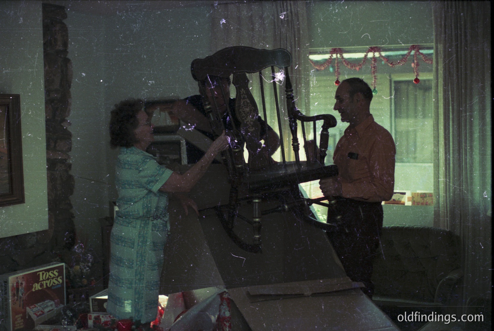 Vintage indoor scene featuring a woman in a striped dress and a man in a military uniform playing a vintage upright piano. Decor includes red paper garlands and a "Toss Across" box on a table. Mid-20th century domestic setting, likely 1950s–1960s.