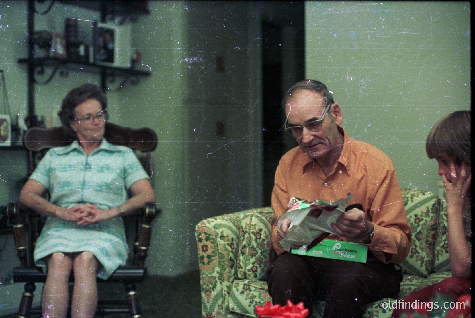Vintage indoor portrait featuring three adults in a living room setting, likely mid-20th century. The man in an orange shirt reads a magazine, while the woman in a teal dress sits beside him. A child in a floral dress sits on the right. Floral-patterned armchairs and a shelf with decorative items fill the background.