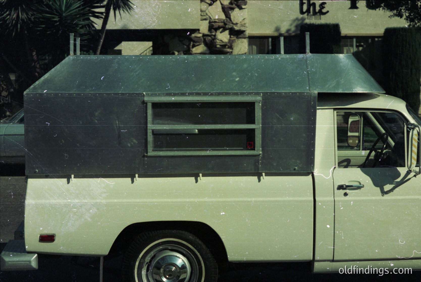 Vintage delivery van with boxy, utilitarian design—distinctive flat roof, rear sliding window, and minimalist exterior. Likely a 1970s–1980s model, possibly for commercial or postal use. Urban setting with partial signage ("the...") and palm trees in background.