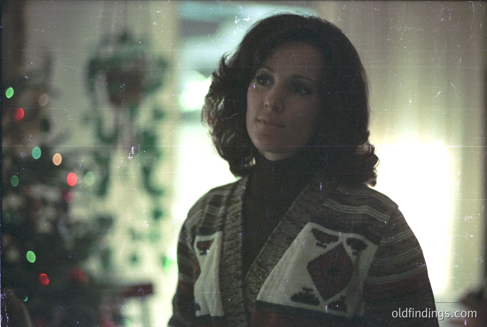 Vintage portrait of a woman in a 1970s-style sweater with geometric patterns, standing near a decorated Christmas tree. Soft focus and warm lighting evoke nostalgic holiday ambiance.