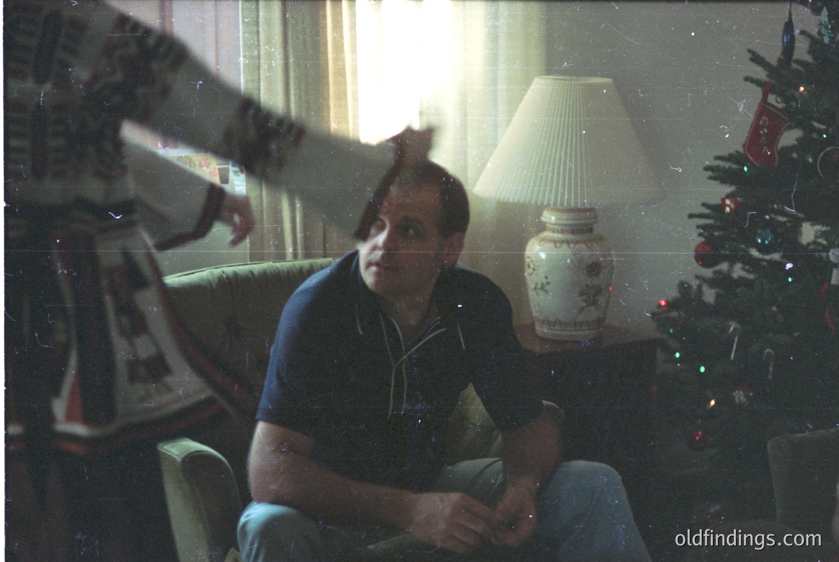 Vintage indoor portrait of a man seated on a mid-century armchair, holding a cigarette. Decor includes a decorated Christmas tree with red/white ornaments, a floral-patterned lamp, and a framed photo on the wall. Warm, nostalgic lighting suggests holiday season, likely 1960s–1970s.