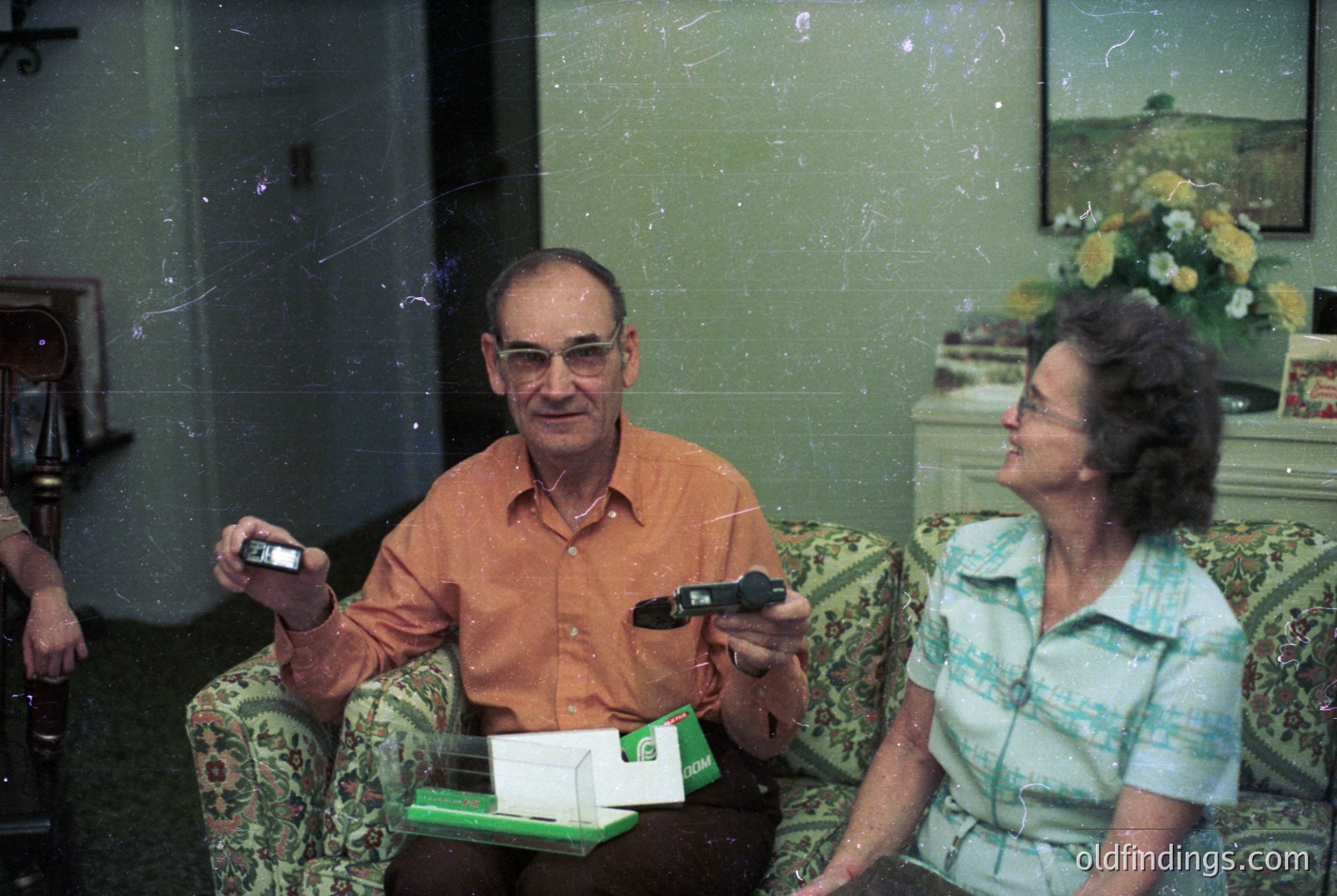 Vintage indoor portrait of two elderly adults in mid-20th-century clothing, likely 1970s–1980s. Man in an orange shirt holds a portable cassette player; woman in a patterned blouse sits beside him. Green floral-patterned sofa and framed artwork in background suggest a mid-century home setting. Warm, nostalgic lighting enhances vintage aesthetic.